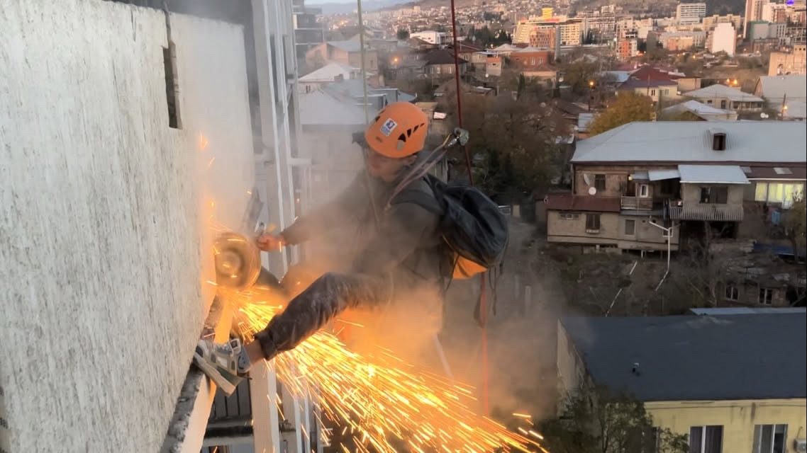 ალპინისტი ფასადს ჭრის, ირგვლივ ნაპერწკლები, ფონზე ქალაქი